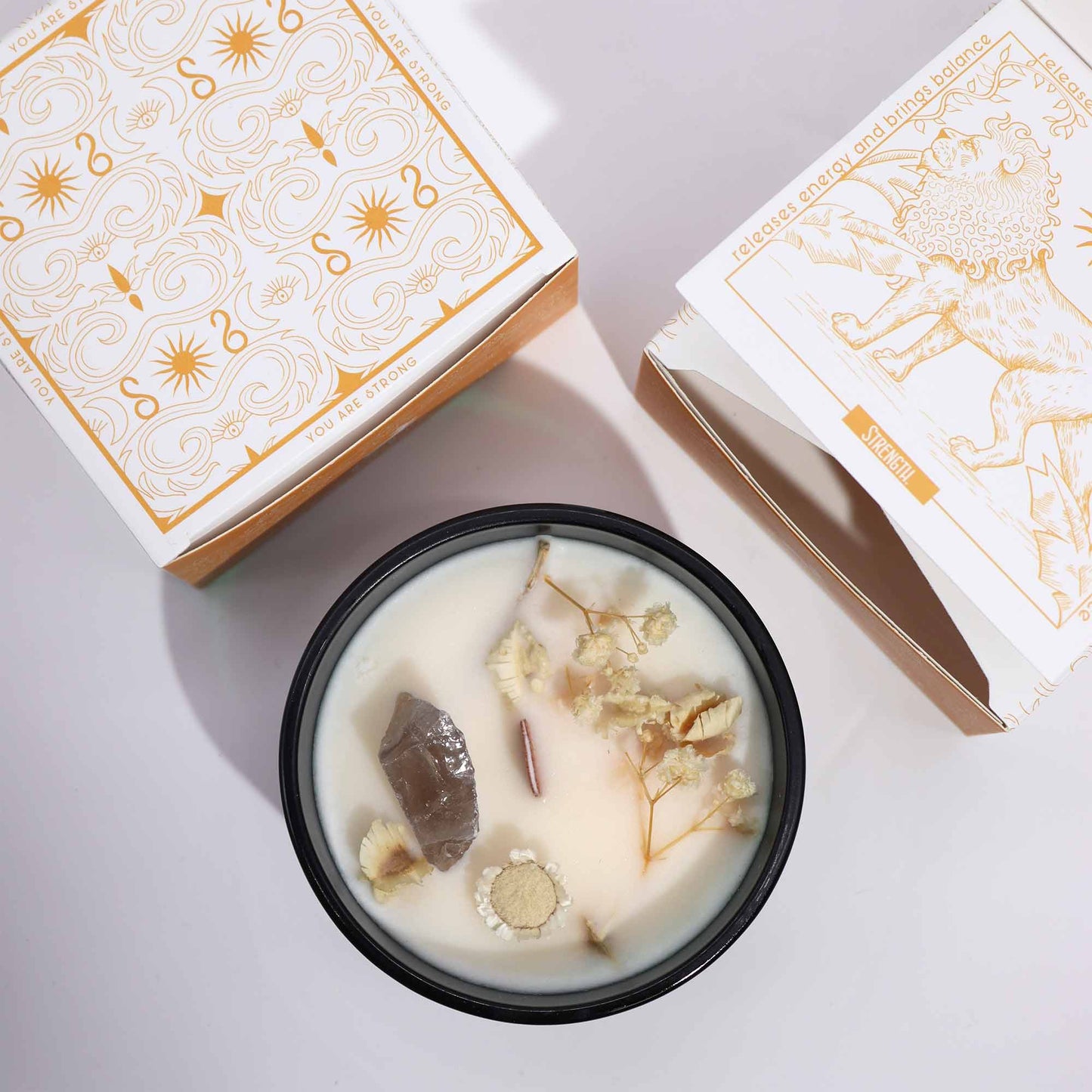 Crystal infused flower candle with dried flowers and crystal in a black container beside decorative packaging