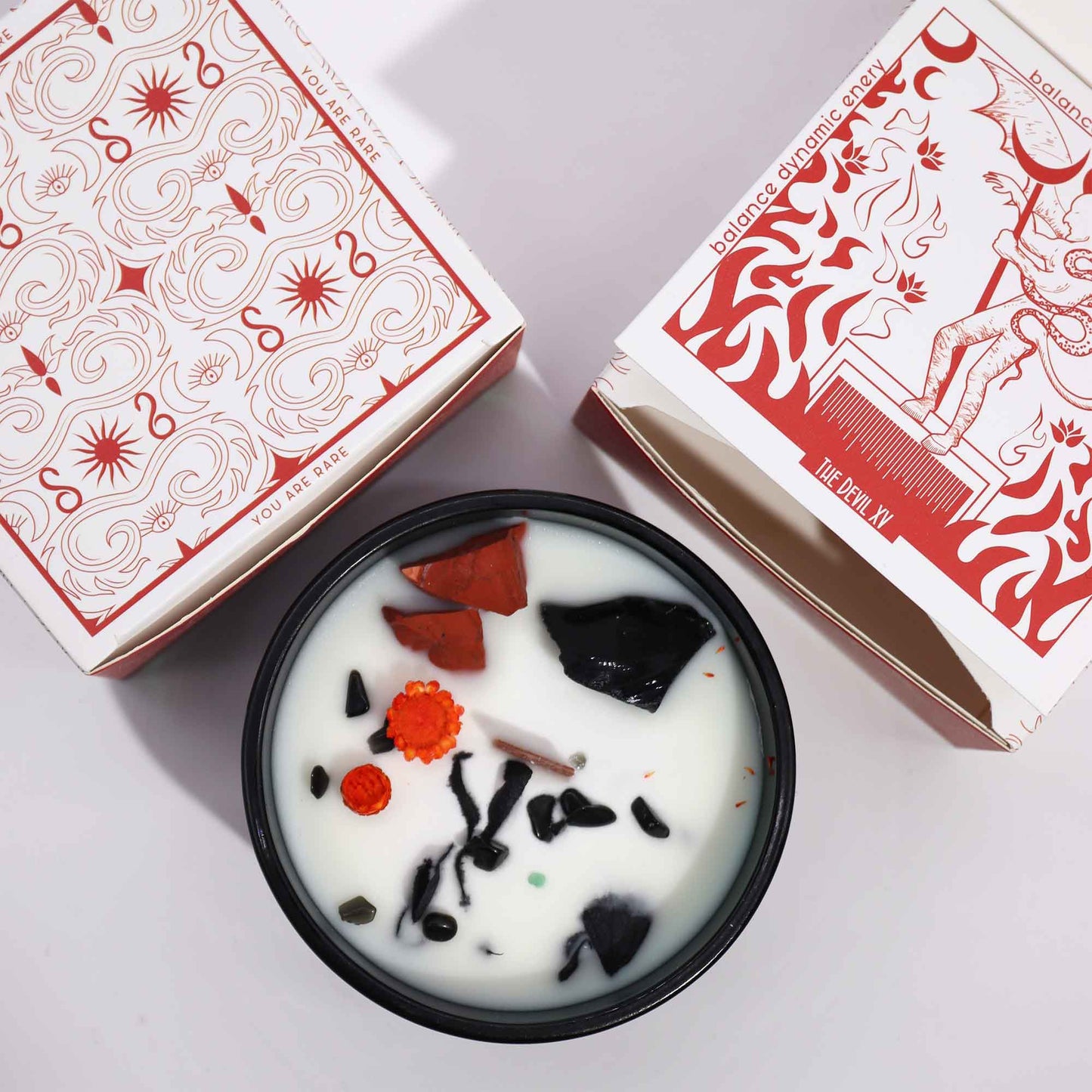 Crystal infused candle with black and orange stones in black container alongside decorative white and red packaging