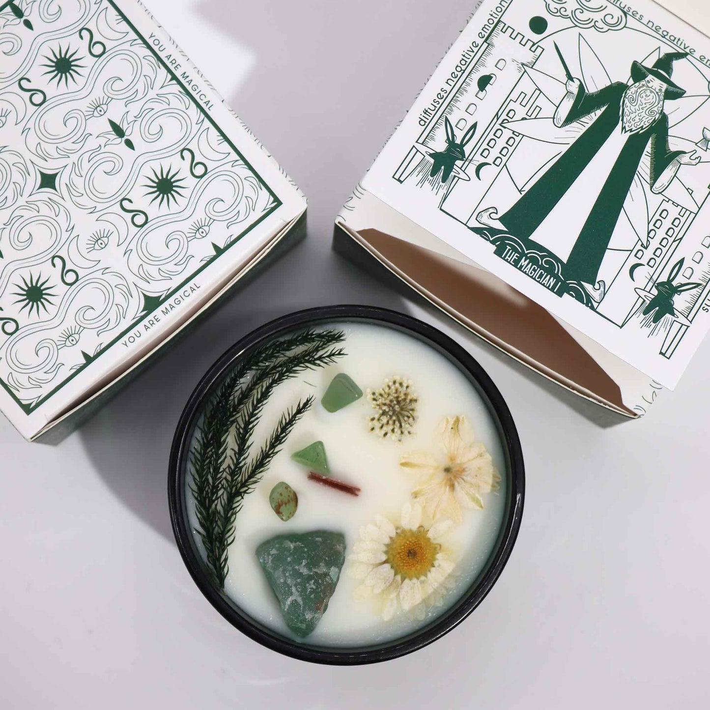 Crystal infused flower candle with green crystals and dried flowers in decorative black container beside mystical-themed packaging