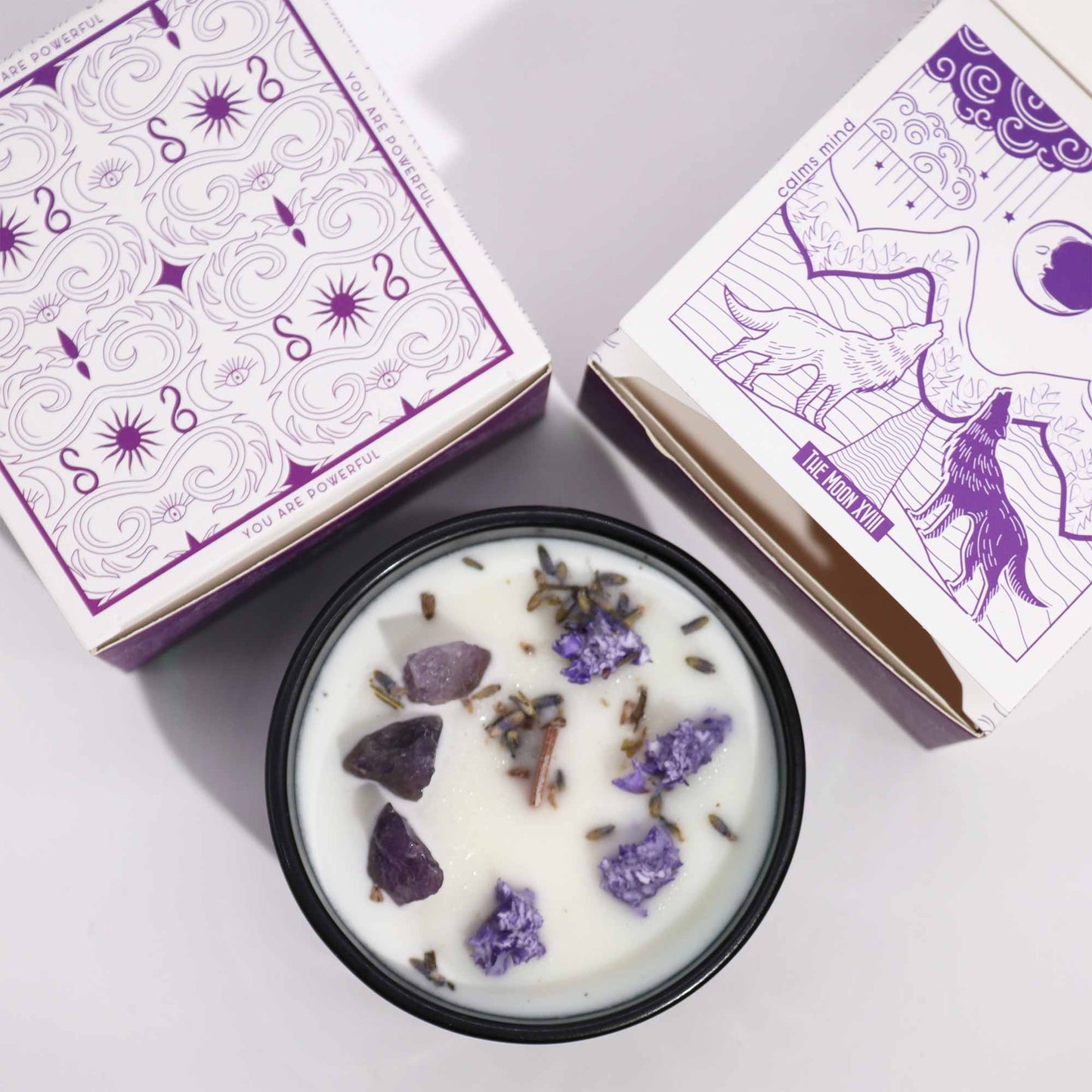 Crystal infused flower candle with purple stones and dried flowers in black container beside decorative purple and white packaging
